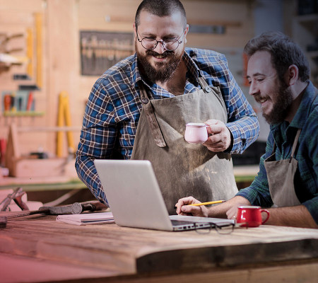 Zwei Männer mit Bart und in Arbeitskleidung stehen gut gelaunt in einer Werkstatt und blicken auf einen geöffneten Laptop.