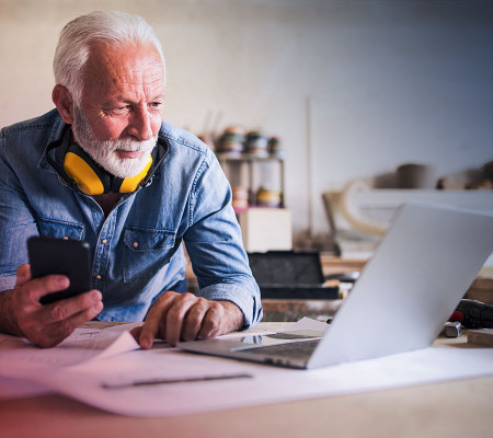 Ein älterer Mann mit lichtem Haar und weißem Bart steht in der Werkstatt. Er hält ein Smartphone in seiner Hand und blickt auf einen geöffneten Laptop.